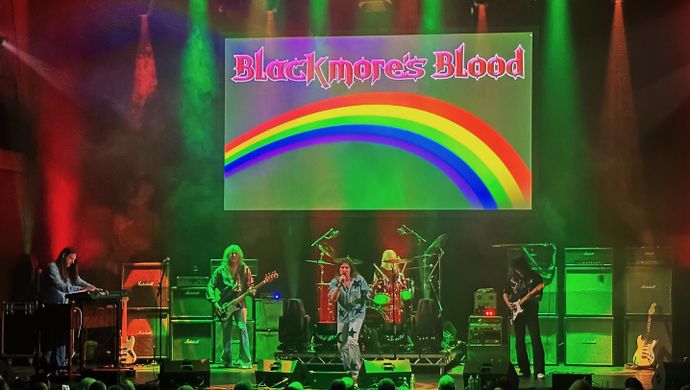 A photographic depiction of the band Blackmore's Blood engaged in a live performance before a graphical representation of a rainbow.  the lead singer is wearing denim garments emblazoned with a crescent moon and various stars. 