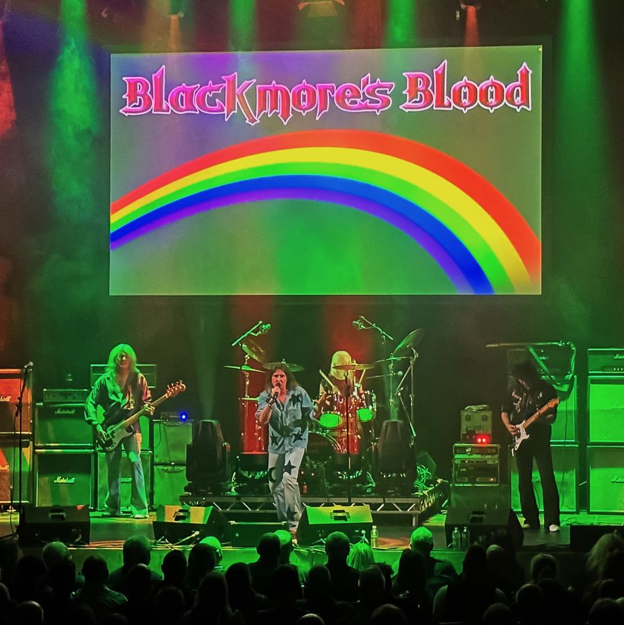 A photographic depiction of the band Blackmore's Blood engaged in a live performance before a graphical representation of a rainbow.  the lead singer is wearing denim garments emblazoned with a crescent moon and various stars. 